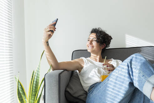 Smiling young woman sitting on couch with a soft drink taking a selfie - JPTF00233