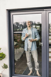 Man standing by glass door, drinking coffee, smiling - UUF18039