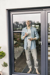 Mann steht an einer Glastür, trinkt Kaffee, lächelt - UUF18039