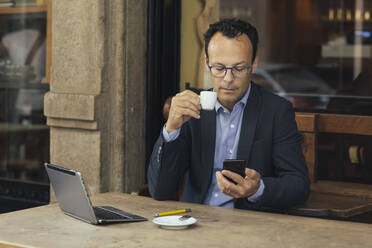Geschäftsmann mit Laptop in einem Café, der auf sein Smartphone schaut - ALBF00887