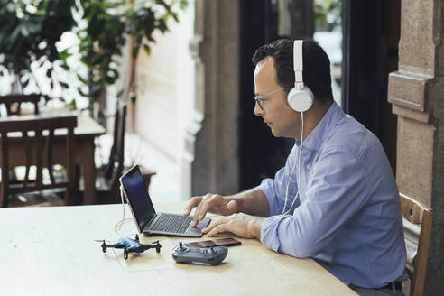 Businessman with headphones, laptop and drone in a coffee shop - ALBF00889