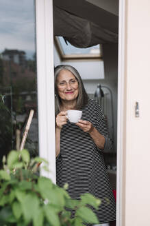 Porträt einer glücklichen älteren Frau, die mit einem Schluck Kaffee an der geöffneten Balkontür lehnt und in die Ferne schaut - ALBF00897