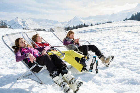 Drei jugendliche Skifahrerinnen sitzen in Liegestühlen in einer verschneiten Landschaft, Tirol, Steiermark, Österreich - CUF51643