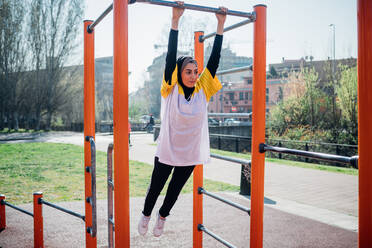 Calisthenics class at outdoor gym, young woman swinging from exercise equipment - CUF51687