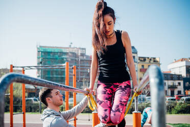 Calisthenics class at outdoor gym, male trainer encouraging young woman on parallel bars - CUF51701