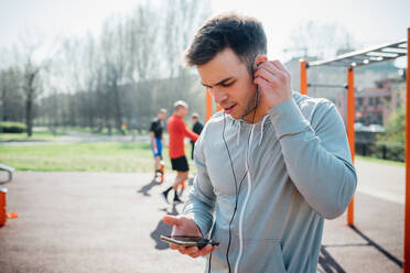 Calisthenics-Kurs in einem Fitnessstudio im Freien, junger Mann setzt Kopfhörer ein - CUF51706