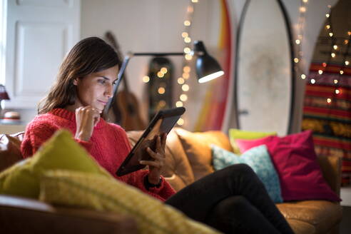 Junge Frau entspannt sich auf dem Wohnzimmersofa und schaut auf ein digitales Tablet - CUF51779