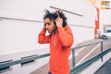 Young man putting on headphones walking past concrete wall - CUF51832