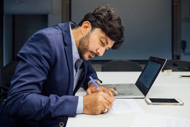 Geschäftsmann schreibt Notizen und benutzt Laptop im Büro - CUF51845