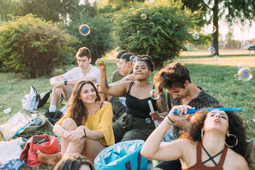 Eine Gruppe von Freunden entspannt sich beim Picknick im Park und bläst Seifenblasen - CUF51898