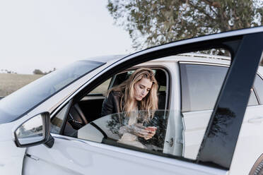 Blonde Frau benutzt Smartphone, sitzt in weißem Auto - ERRF01523