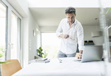 Architect working on blueprint in a bright office, using laptop - UUF18042