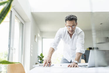 Architect working on blueprint in a bright office, using laptop - UUF18045