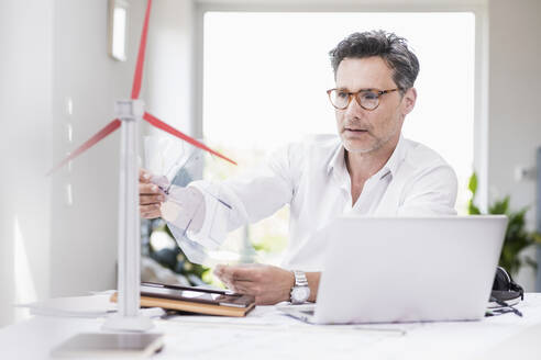 Businessma n sitting in office, using laptop, looking at model of a windwheel - UUF18068