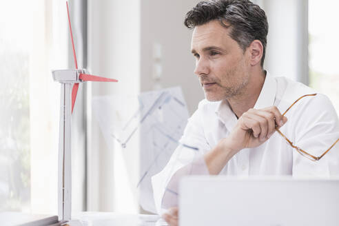 Businessma n sitting in office, using laptop, looking at model of a windwheel - UUF18069