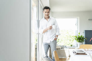 Mature man leaning on window, drinking coffee at home - UUF18082