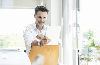 Mature man sitting on chair backwards, using digital tablet - UUF18093