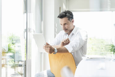 Mature man sitting on chair backwards, using digital tablet - UUF18094