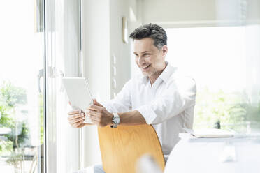 Mature man sitting on chair backwards, using digital tablet - UUF18096