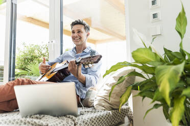 Mature man practising on the guitar, using laptop and earphones - UUF18098