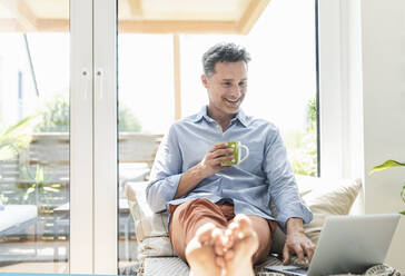 Mature man sitting on couch, using laptop, drinking coffee - UUF18106