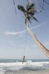 Frau schwingt sich in voller Länge von einer Palme am Strand gegen den Himmel, Sri Lanka - LHPF00717