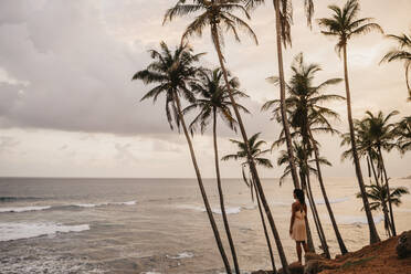 Woman looking at sea while standing by palm trees against cloudy sky, Sri Lanka - LHPF00722