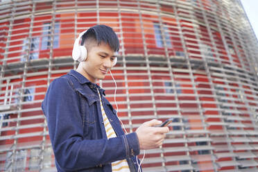 Porträt eines lächelnden jungen Mannes mit Kopfhörern, der auf sein Mobiltelefon schaut, Barcelona, Spanien - DVGF00034