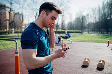 Gymnastik im Freien, junger Mann hört Kopfhörer und schaut auf sein Smartphone - CUF51966
