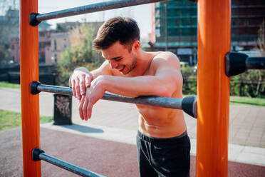 Calisthenics at outdoor gym, bare chested young man leaning on exercise equipment exhausted - CUF51975