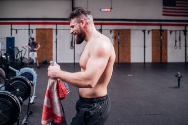 Junger Mann mit nacktem Oberkörper beim Training, hält Wasserflasche und Handtuch im Fitnessstudio, Seitenansicht - CUF52030