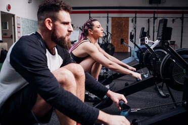 Junge Frau und Mann trainieren gemeinsam an Rudergeräten in einem Fitnessstudio, Seitenansicht - CUF52043