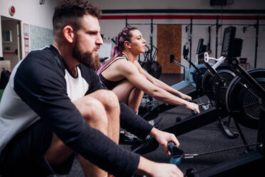 Young woman and man training on rowing machines together in gym, side view - CUF52043