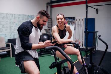 Young woman and man training together on gym exercise bikes, looking at smartphone - CUF52044