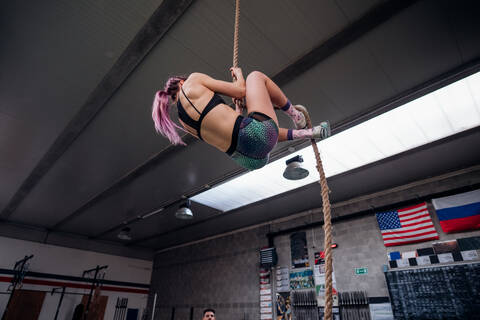 Junge Frau trainiert, klettert an einem Übungsseil in einem Fitnessstudio hoch, niedriger Blickwinkel, lizenzfreies Stockfoto