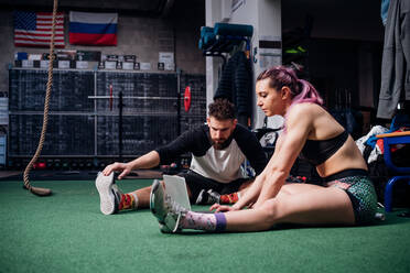 Junge Frau und Mann trainieren zusammen im Fitnessstudio, sitzen auf dem Boden und machen Aufwärmübungen und schauen auf den Laptop - CUF52055