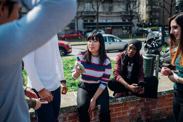 Junge weibliche und männliche erwachsene Freunde sitzen an der Wand, unterhalten sich und schauen auf ihre Smartphones - CUF52076
