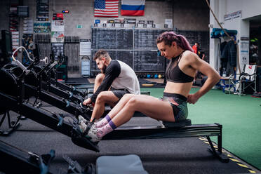 Young woman and man training together, preparing to use rowing machines in gym - CUF52088