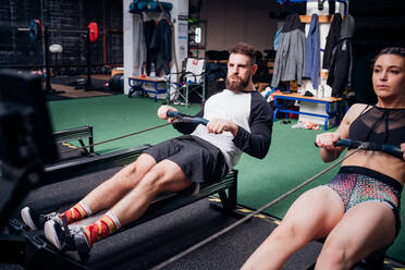 Young woman and man training on rowing machines together in gym - CUF52091