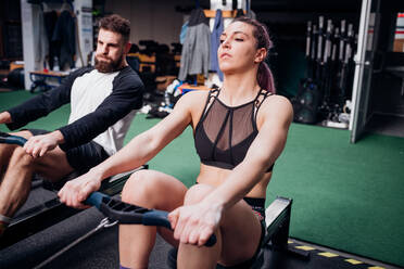 Young woman and man training on rowing machines together in gym - CUF52092