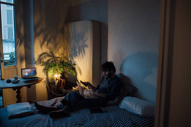 Bärtiger junger Mann mit Smartphone und Laptop im Bett - CUF52121