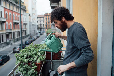 Bärtiger junger Mann gießt Pflanzen auf dem Balkon - CUF52126