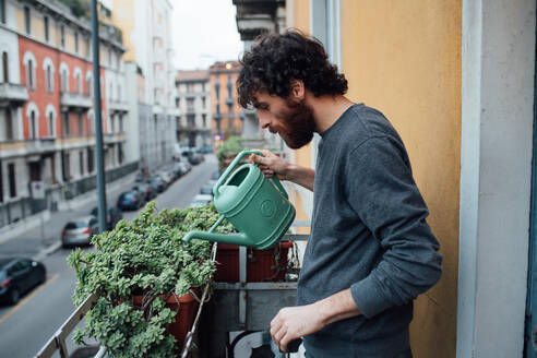 Bärtiger junger Mann gießt Pflanzen auf dem Balkon - CUF52126