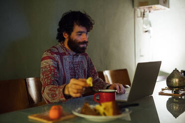 Bärtiger junger Mann beim Frühstück, der zu Hause einen Laptop benutzt - CUF52141