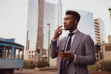 Businessman using smartphone, smoking on pavement, Milano, Lombardia, Italy - CUF52181