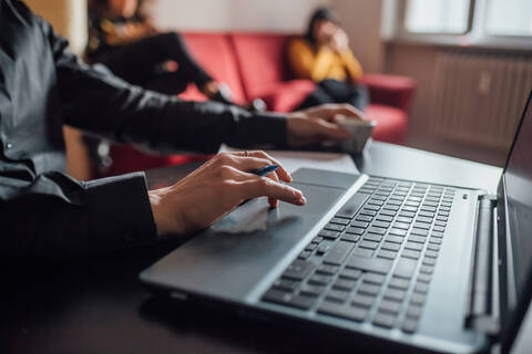 Mann mit Laptop, Freunde entspannen sich im Hintergrund im Heimbüro, lizenzfreies Stockfoto