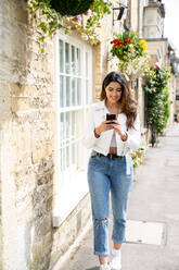 Junge Frau schlendert die Dorfstraße entlang und schaut auf ihr Smartphone, Cotswolds, England - CUF52449