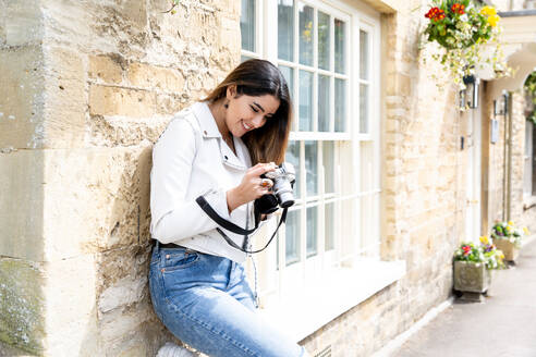 Junge Frau lehnt an einer Hauswand und betrachtet Fotos auf einer Digitalkamera, Cotswolds, England - CUF52451