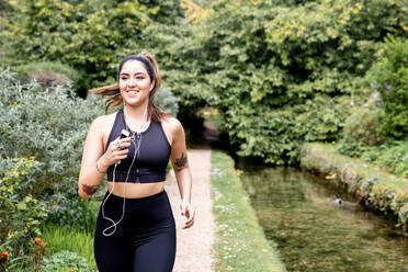 Young female runner listening to earphones while running on riverside path - CUF52456