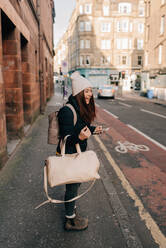 Woman waiting on kerb with cellphone and luggage, Edinburgh, Scotland - CUF52492